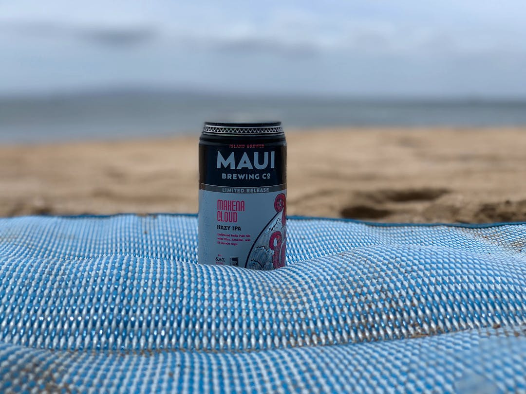 Shot of a can of Maui Brewing Co beer, on a blanket, with the beach in the background.